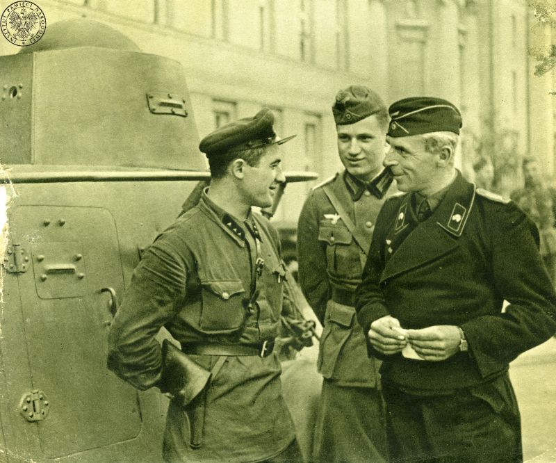 Three soldiers - one Soviet, two German - are talking to each other. They are smiling. A tank in the background.