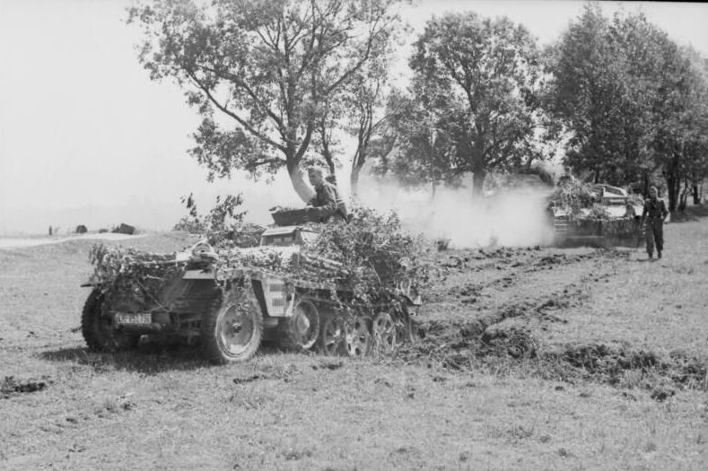 Combat vehicles masked with branches driving along a country road. Trees in the background.