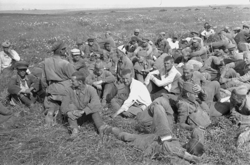 A group of soldiers is sitting on a meadow, some are lying down. Some have their uniforms incomplete.