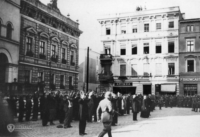 Town buildings. Probably the market square. Men are standing in front of the entrance to the building. They have their hands raised. Next to them a large group of soldiers.