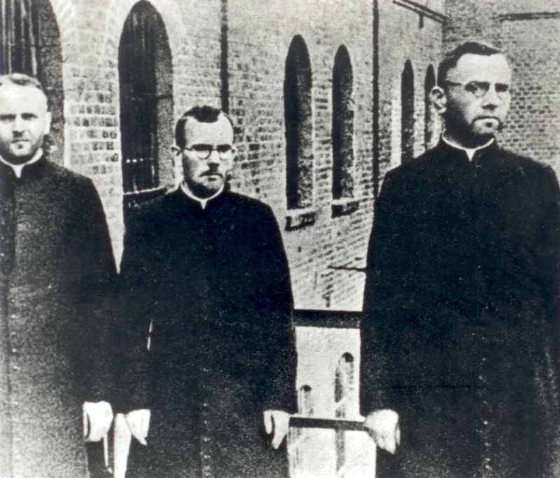 Four priests in habits standing in front of the entrance to the building. Their faces are directed towards the building, concentrated with serious expressions.