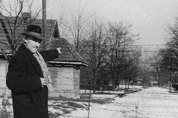 A tall, elegant man in coat and hat pointing with his hand towards the road. Buildings on the left, a barbed wire fence behind the man.