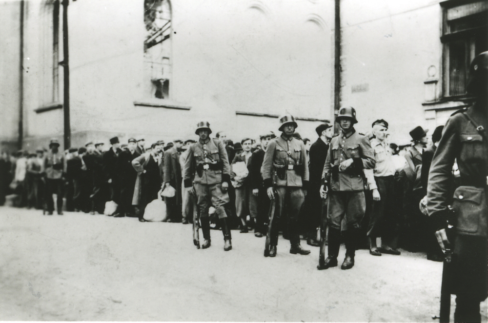 In the foreground, German soldiers with rifles. Behind them the prisoners of the first transport to Auschwitz standing in a column.