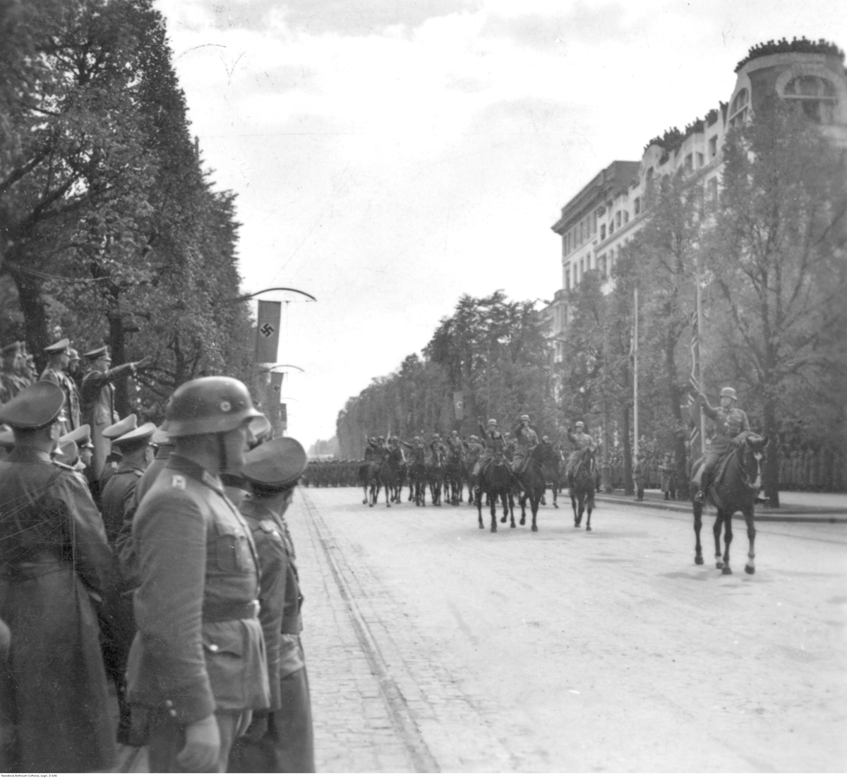 Street of Warsaw. There is a parade in the middle. At its front, a German soldier riding on horseback and saluting Hitler. On the left, German soldiers and Adolf Hitler saluting.