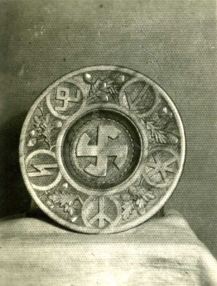 Round, decorated, wooden plate. Round swastika in the middle. Five symbols on the sides - Germanic runes, plant motifs between them: oak leaves and acorns.