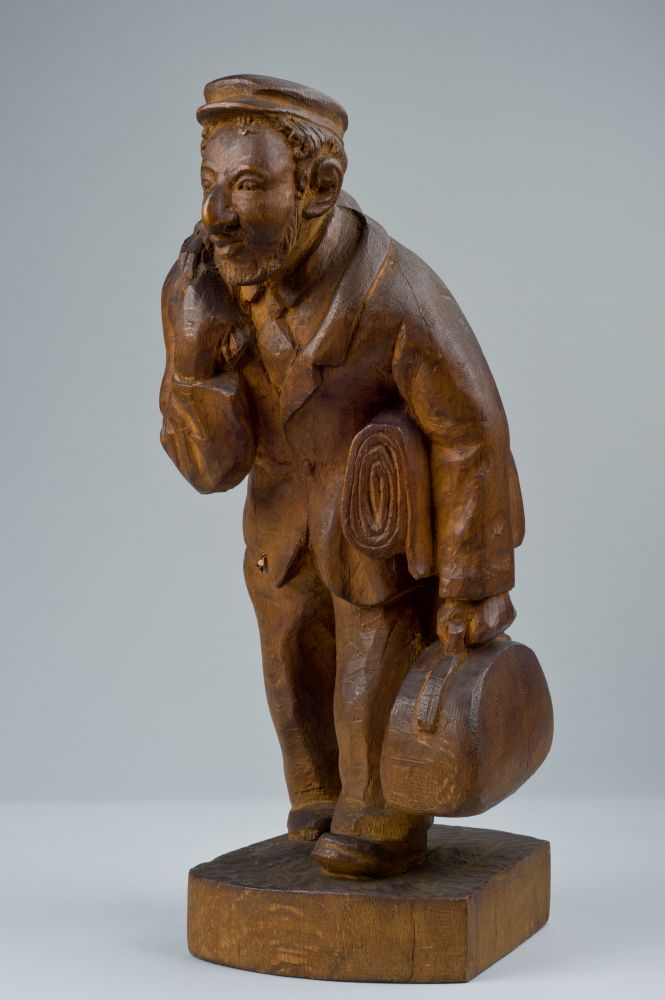 Wooden figure of a man. Round face, facial hair. Prominent, slightly curved nose. A flat cap on his head. He is holding a bag in his hand and a blanket under his arm. In his other hand he is holding a bag slung over his shoulder.