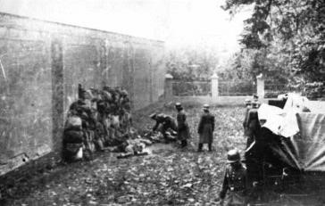 A high wall, people are standing prepared to be executed. Some have already been shot - the bodies are lying motionless next to the standing people. German soldiers in front of them. On the right a car with a tarpaulin, behind it stands the execution platoon.