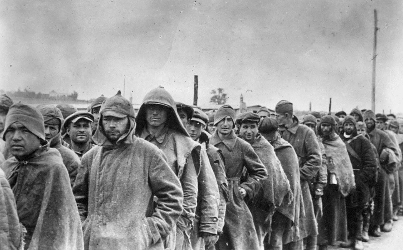 A column of Soviet prisoners heading in an unknown direction. They are dressed in Soviet uniforms, long coats, and torn blankets. Some have characteristic pointy hats on their heads.