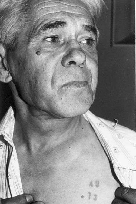 An elderly man showing a number tattooed the left side of his chest.  Calm expression, gazing beyond the camera lens.
