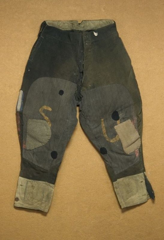 Torn, tattered trousers. On the left leg the letters SU written with yellow paint and a red painted stripe along the leg.

