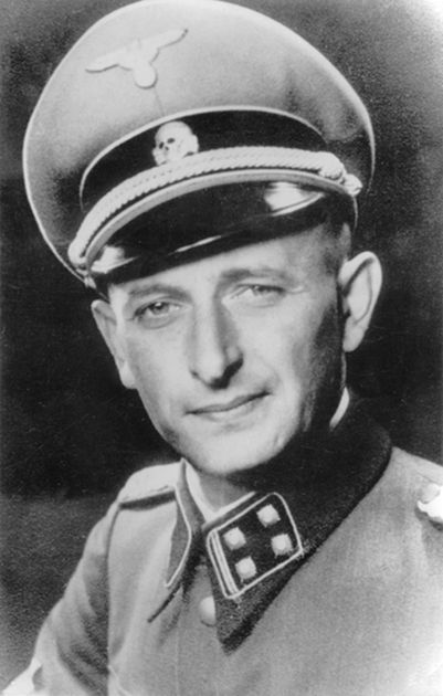 Adolf Eichmann. A man in an SS uniform with an eagle and skull insignia on his cap. He looks directly at the camera, smiling.