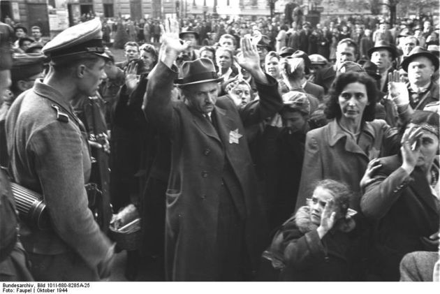 Crowd of people of all ages. In the foreground a man wearing a hat with his hands raised, next to him a girl has her hands raised. They are looking at a German soldier who is talking to them.