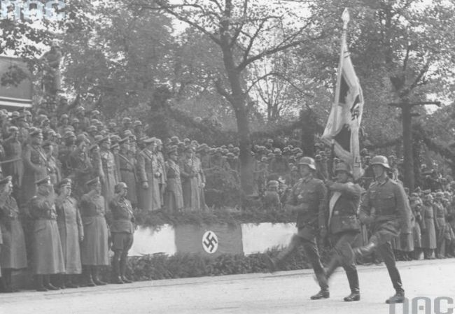 German soldiers saluting while standing on a stand of honour with a swastika on it.