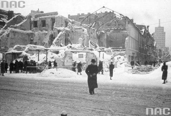 In the foreground a man standing, wearing a hat and a coat; further away groups of passers-by with the ruins of a tenement building visible in the background.