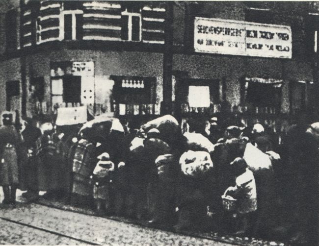 A crowd of people wrapped in blankets with small children standing by the tram tracks. Everyone has their belongings kept in bundles. Next to them an SS man guarding the crowd.
