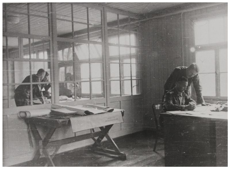 Camp construction office. A man is sitting at a table, another is standing above him. They are leaning over sheets of paper. There is a glass partition in the middle of the room. Under it, there is a table covered with papers, plans and documents.