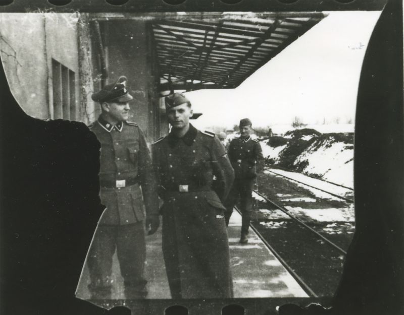 Three SS men in uniforms standing in front of the SS-Unterkunftsgebäude building. Railway tracks on the right. Winter, snow.