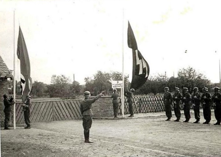 SS men in uniforms, with helmets on their heads, standing in a row and presenting weapons. One is saluting. Four men are hanging two SS flags on both sides of the wooden gate. On the right, an inscription above the gate: Lazaret Auschwitz.