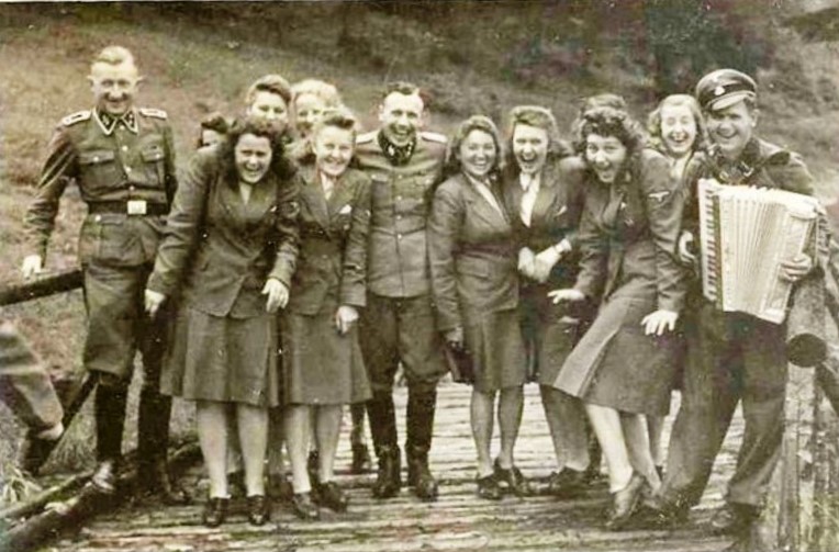 Women from the SS women's auxiliary formations in uniforms and three SS men in uniforms during their stay at the recreation center for SS men in Międzybrodzie (SS-Sola Hütte). They are standing on a wooden bridge, laughing, and one of the SS men is playing the accordion.