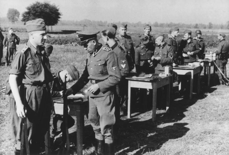 SS men in uniforms with rifles at a field shooting range. They are gathering in groups around tables with packages of rifle ammunition and documents. Two of them are writing something down.