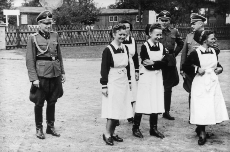Four women - German Red Cross nurses in white aprons and three SS men in uniforms. They are standing in the square, a wooden fence and buildings behind them. They are laughing.