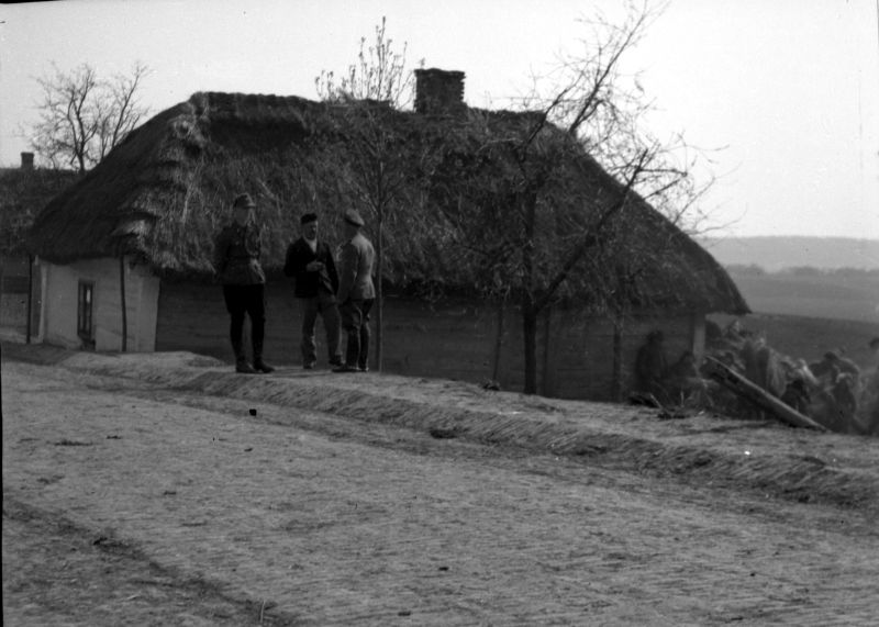 A thatched cottage. In front of the thatched cottage, three men stand in front of the road and talk.