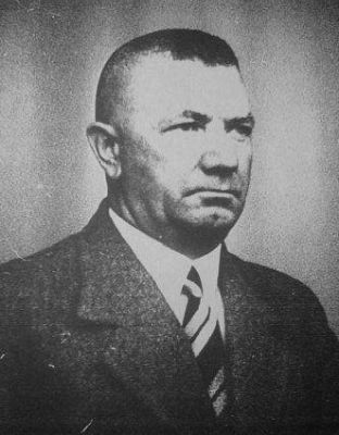 A man in a suit. Serious face, fierce expression, short hair. Face turned slightly to the right side.