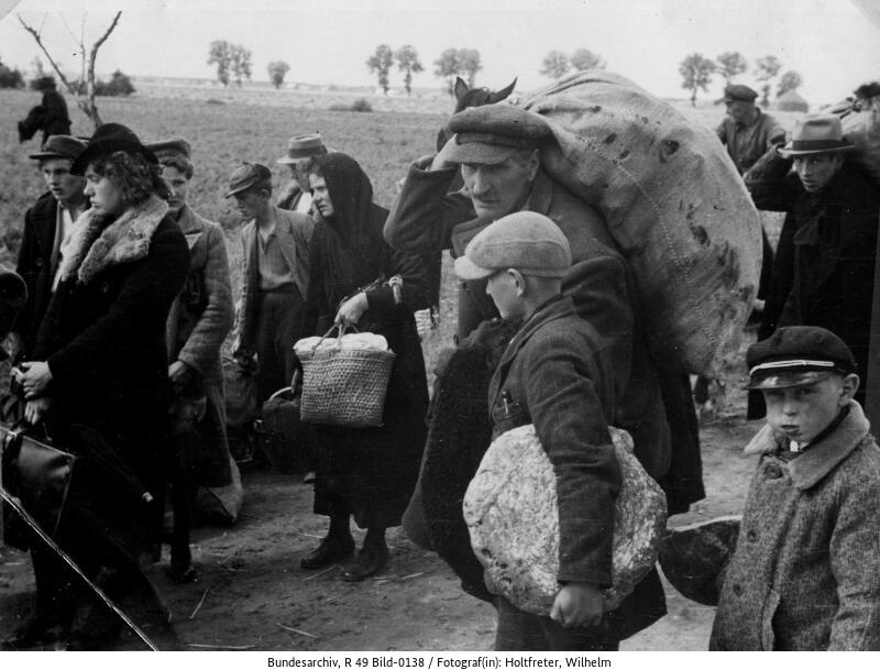 A crowd of people - men, women and children with bundles follow the gravel road.