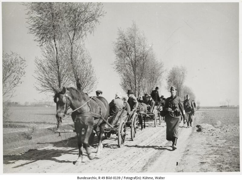 Crowds of civilians on the gravel road. Horse-drawn carts harnessed to the road. On the right side German soldiers.