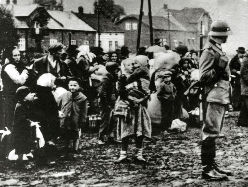The moment of displacement of the inhabitants of the Zamość region. Women, children, men, carts with belongings. Rural buildings in the background. On the right side, SS man standing sideways. Hands folded behind his back, observing.