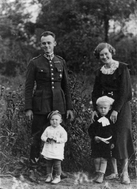 A man in a Polish army uniform, smiling, standing in an orchard. Next to him is a woman, smiling, wearing a dark dress. Children standing in front of the couple: a younger boy, about two years old, a 4–5-year-old girl.