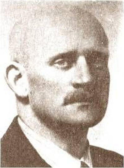 Portrait photograph of a middle-aged man. Face focused, serious, gaze directed at the lens. Dressed in a jacket, a shirt and a tie. Bald, short-cropped moustache.