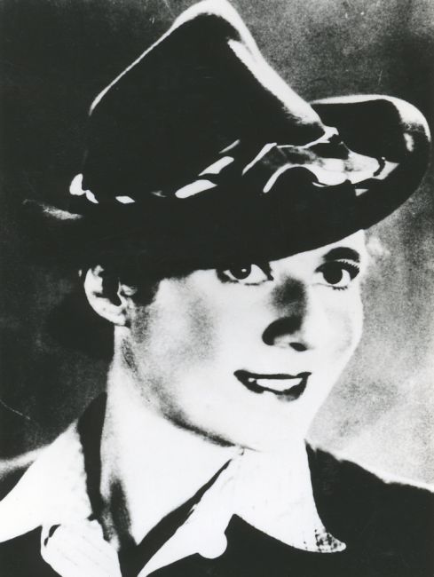 Portrait photograph of a young woman wearing a decorated hat. Smiling, face and gaze turned slightly to the right. Shirt collar turned up over the top of the jacket. Light hair.