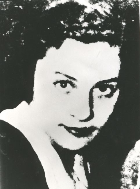 Portrait photograph of a young woman. Face and gaze turned towards the lens. Dark hair decoratively styled upwards. Shirt collar turned up over the top of the jacket.